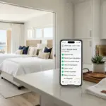 Smartphone displaying STR cleaning management app on a vacation rental kitchen counter with freshly made beds in background